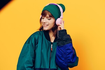 horizontal photo of a beautiful woman in a jacket and hat listening to music with pink headphones holding them with her hands