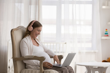 Pretty pregnant woman in headphones sit on armchair with laptop, smile enjoy video call with...