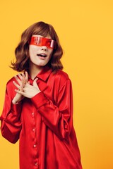 an attractive, sweet, thoughtful woman stands on a yellow background in a red shirt and fashionable glasses of an interesting shape, holding her fingers folded together near her chest
