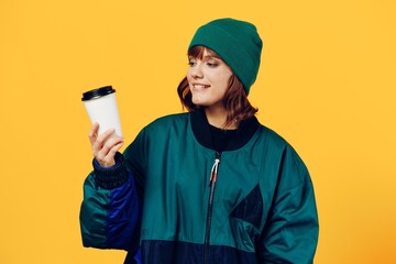 Fototapeta na wymiar a happy, joyful woman stands on a yellow background in a stylish jacket and holding a coffee glass in her hands smiles pleasantly looking at him