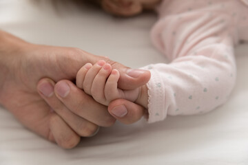Close up cropped shot of tiny arm of cute newborn, baby girl holds daddy thumb during carefree healthy sleep in bed. Family bond and ties, parent unconditional love, cherish, happy paternity concept