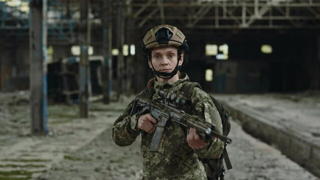 Professionally trained female soldier walking at destroyed plant and using military rifle for looking in sight. Armed caucasian woman wearing uniform and helmet.