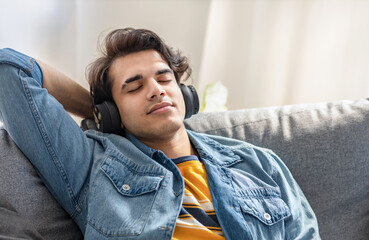 Young caucasian man with headphones resting on sofa at home, listening to music, dreaming, thinking positively