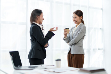 Two beautiful Asian business women standing chatting during a break relaxing coffee, discussing work and analyzing data at work.