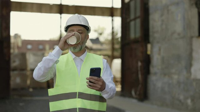 Caucasian mature foreman browsing internet on modern smartphone and drinking hot coffee-to-go while walking at construction site. Bearded architect in helmet and reflective vest having break at work.