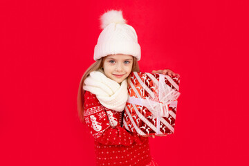 a child girl in a winter hat and sweater with gifts on a red monochrome isolated background rejoices and smiles, the concept of new year and Christmas, space for text