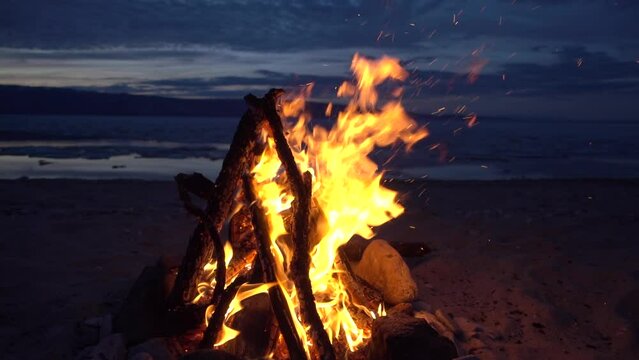 Romantic bonfire balefire at night after sunset burns on shore of lake, sea. Spring. Firewood, fire. Travel, tourist landscape details, sparks fly. Atmospheric cozy stock movie, Gimbal, slow motion