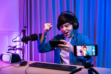 Playing video games on smartphone. Young asian handsome man sitting on chair holding cellphone in his hand. Exited streamer wearing headset  in neon room .Esport streaming game online.