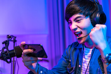 Playing video games on smartphone. Young asian handsome man sitting on chair holding cellphone in his hand. Exited streamer wearing headset  in neon room .Esport streaming game online.