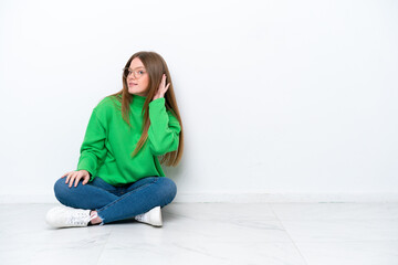 Young caucasian woman sitting on the floor isolated on white background listening to something by putting hand on the ear - obrazy, fototapety, plakaty