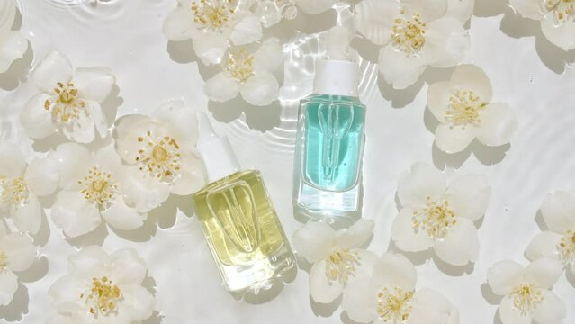 Cosmetic bottle, vial and jasmine petals flowers on water surface with drops. Pure water with reflections sunlight and shadows. Slow motion of waves water. Design, advertising, products. 4k