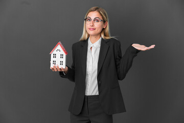 Blonde Uruguayan girl holding a house toy isolated on black background having doubts while raising hands