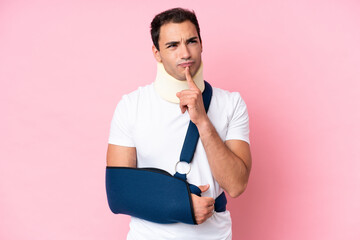 Young caucasian man with broken arm and wearing a sling isolated on pink background having doubts while looking up