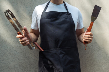 A man wearing black chef's apron, holding barbecue tools: bbq tongs, spatula. - obrazy, fototapety, plakaty