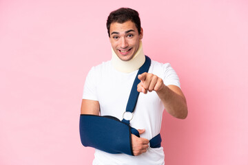 Young caucasian man with broken arm and wearing a sling isolated on pink background surprised and...