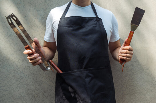 A man wearing black chef's apron, holding barbecue tools: bbq tongs, spatula.