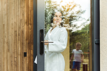 Young woman dressed in white bathrobe leaving the modern house on the terrace, looking after her child through the glass of the door, enjoying beautiful outdoors atmosphere. - obrazy, fototapety, plakaty
