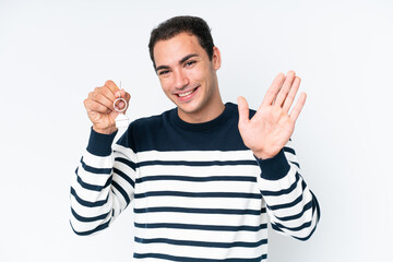 Naklejka premium Young caucasian man holding home keys isolated on white background saluting with hand with happy expression