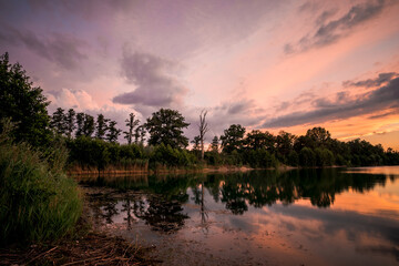 Obraz premium Epic red and gold clouds over forest lake at sunset. Dramatic cloud cover. Symmetrical reflections on the water, natural mirror. idyllic landscape.