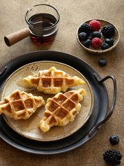 Croffles (combination of croissant and waffle) served in ceramic plate on a retro metal tray. Glass gravy boat with maple syrup and small bowl with with fresh berries. Food trend from South Asia