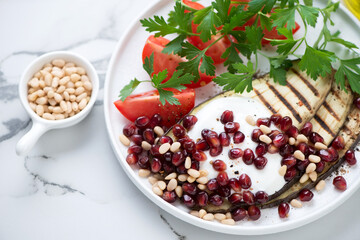 White plate with grilled slices of aubergine, pomegranate seeds, pine nuts, yogurt, tomatoes and parsley, middle close-up