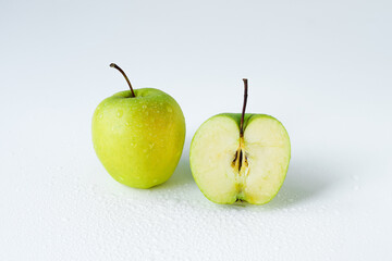 One whole apple and one in a section on a plain light surface. Two round yellow-green apples covered with water drops on the table