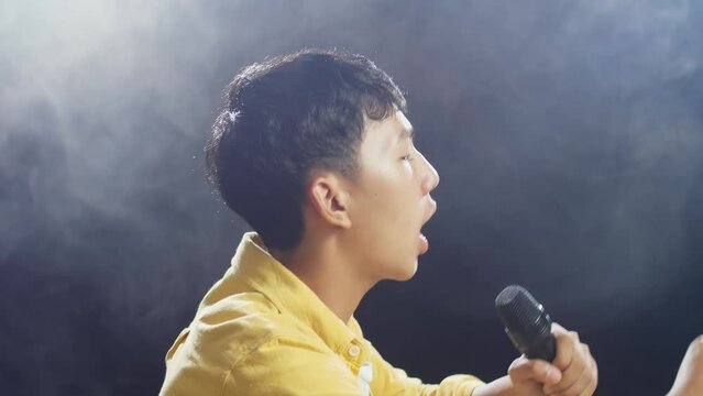 Close Up Side View Of Young Asian Boy Holding A Microphone And Rapping On The White Smoke Black Background
