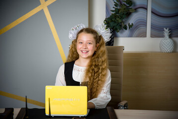 diligent schoolgirl in uniform sitting at the desk with yellow laptop and studying online at school	