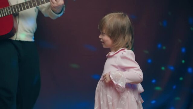 Little girl on stage in pink vintage dress, she dances, her father plays acoustic guitar. Color music is shining. Performance on stage. Early development of children