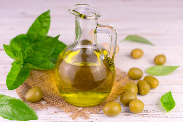 Organic olive oil in a glass bottle and green olives with basil leaves on a white wooden background.
