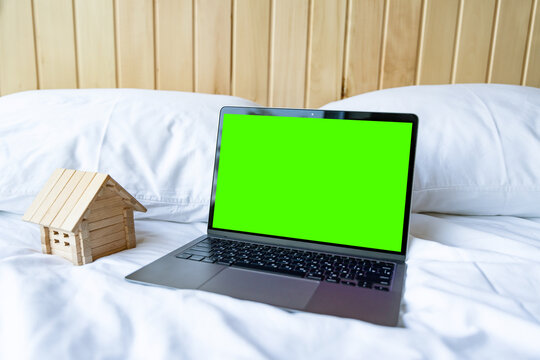 Blank laptop with empty screen and little wooden house lying on the white bed.