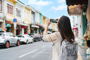 Beautiful young Asian traveler woman using mobile phone take a photo while walking on the city street. Travelling in Phuket Old Town Thailand. Travel concept - obrazy, fototapety, plakaty