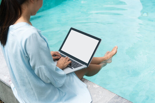 Freelancer using laptop working remotely near swimming pool. Young Asian traveler woman working on computer during her summer holiday. Technology and lifestyle concept. Closeup, copy space