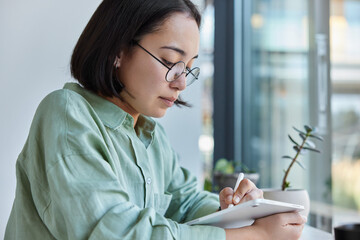 Sideways shot of serious Asian woman focused at tablet screen draws on graphic tablet using stylus...