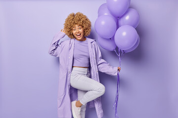 Positive glad woman stands on one leg with bunch of balloons dressed in one colored clothes comes on party to celebrate college leaving looks with cheerful expression has festive mood poses indoor