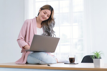 Beautiful young Asian freelancer woman using laptop computer remotely from home. Smiling creative japanese or korean female relax sitting on top desk working communicating online in home office.