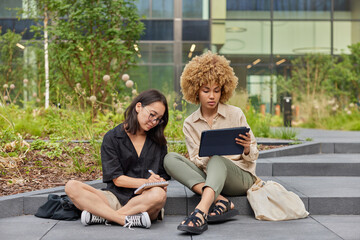 Outdoor shot of two female students of colleague share ideas about coursework make research watch training video via touchpad write down notes in spiral notebook sit on stairs near modern building