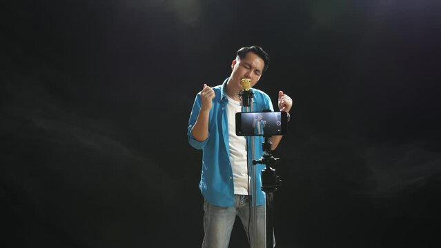 Asian Man Singing Into A Condenser Microphone While Recording Video By Smartphone On The Black Background

