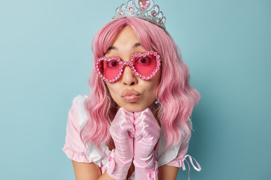 Portrait of lovely pink haired woman wears crown trendy pink sunglasses and gloves keeps hands under chin lips rounded listens something attentively has image of princess or queen dresses for party