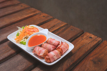 Fried Bacon wrapped sausage rolls with chili sauce and vegetable on wooden table with loft concrete wall in vintage tone style