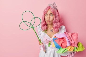 Displeased housewife frowns face looks unhappily away busy doing housework holds basin of colorful laundry and carpet beater wears crown and dress poses against pink background empty space on left