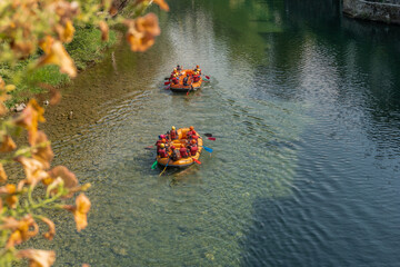 Two groups of people with life jackets, wetsuits, colored paddles and helmets in orange inflatable boats practicing rafting on a calm river without too much current. with copyspace and vegetation