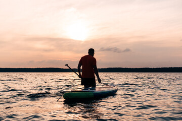 Joyful red bearded man in sunglasses is training on a SUP board on a large lake during sunny day. Stand up paddle boarding - awesome active recreation in nature. 