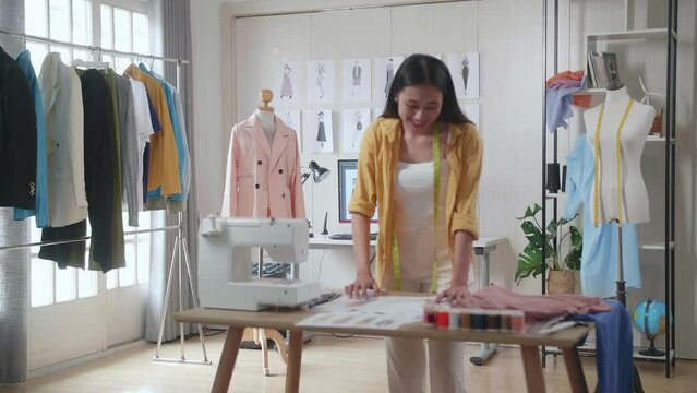 Smiling Asian Female Designer With Sewing Machine Walking To Look At The Pictures On The Wall While Designing Clothes In The Studio

