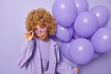 Obraz na płótnie Canvas Stylish teenage girl wears sunglasses and windbreaker keeps lips folded comes on party to celebrate something holds bunch of inflated balloons isolated over purple background. Holiday concept
