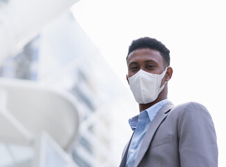 Fototapeta na wymiar businessman wearing mask, standing outside building in city. black working man wearing suit and looking at camera