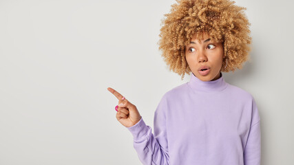 Beautiful surprised young woman with blonde curly hair advertises shocking offer has astonished expression dressed in casual purple jumper isolated over grey background. Omg amazing promo deal