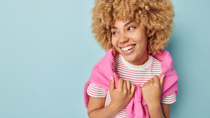 Horizontal shot of curly haired woman smiles broadly looks away wears striped t shirt and pink pullover poses against blue background empty space for your promotional content. Happy emotions