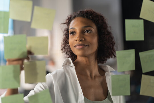 Young African American female employee looking at adhesive sticky notes on glass in the modern office, scrum agile methodology planning project. Shot through the glass