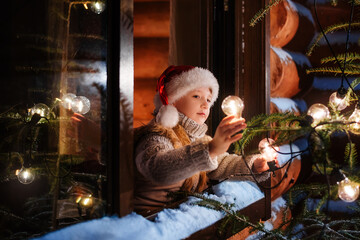 Fototapeta na wymiar A little boy decorates a Christmas tree with a retro garland with warm light bulbs. Holidays winter season. Modern wooden house with large windows.